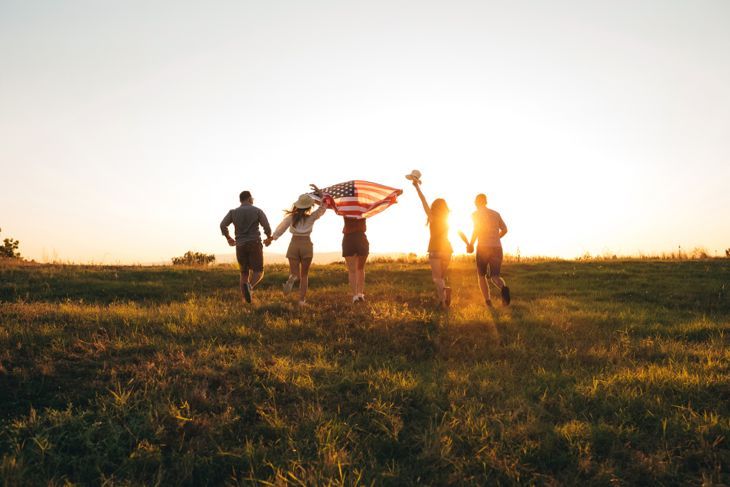 Gruppe junger kaukasischer gutaussehender Leute, die bei Sonnenuntergang die breite amerikanische Flagge ausbreiten.