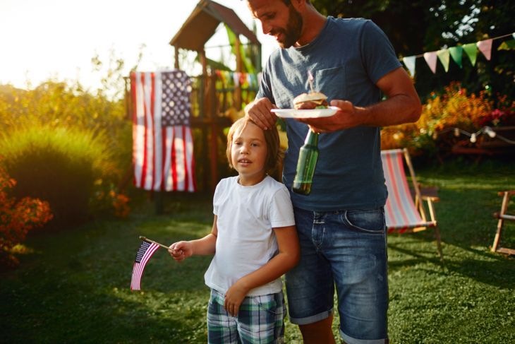 Familie beim Picknick im Hinterhof, der den 4. Juli feiert - Unabhängigkeitstag. Vater hält Burger und sein Sohn hält amerikanische Flagge.